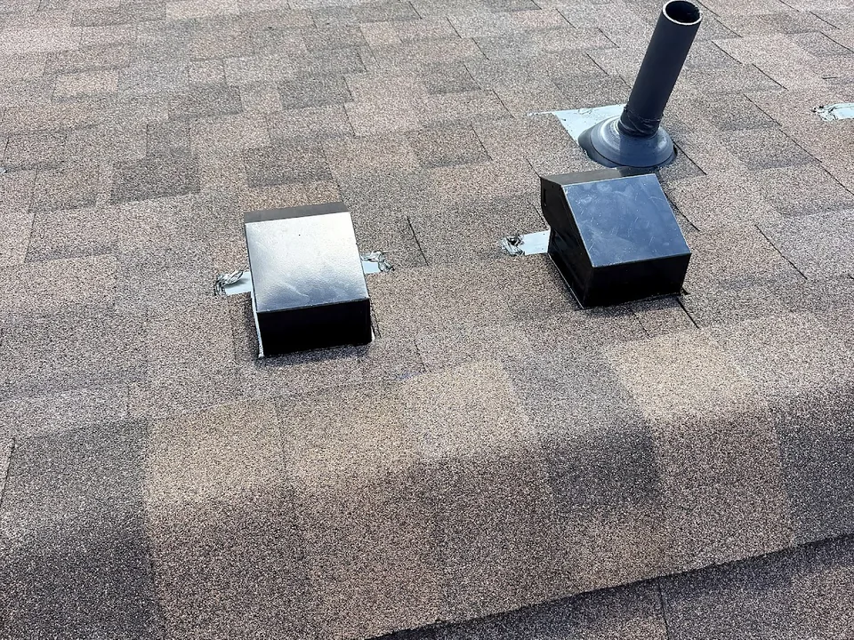Two black box vents and a pipe flashing near the ridge of a brown shingle roof