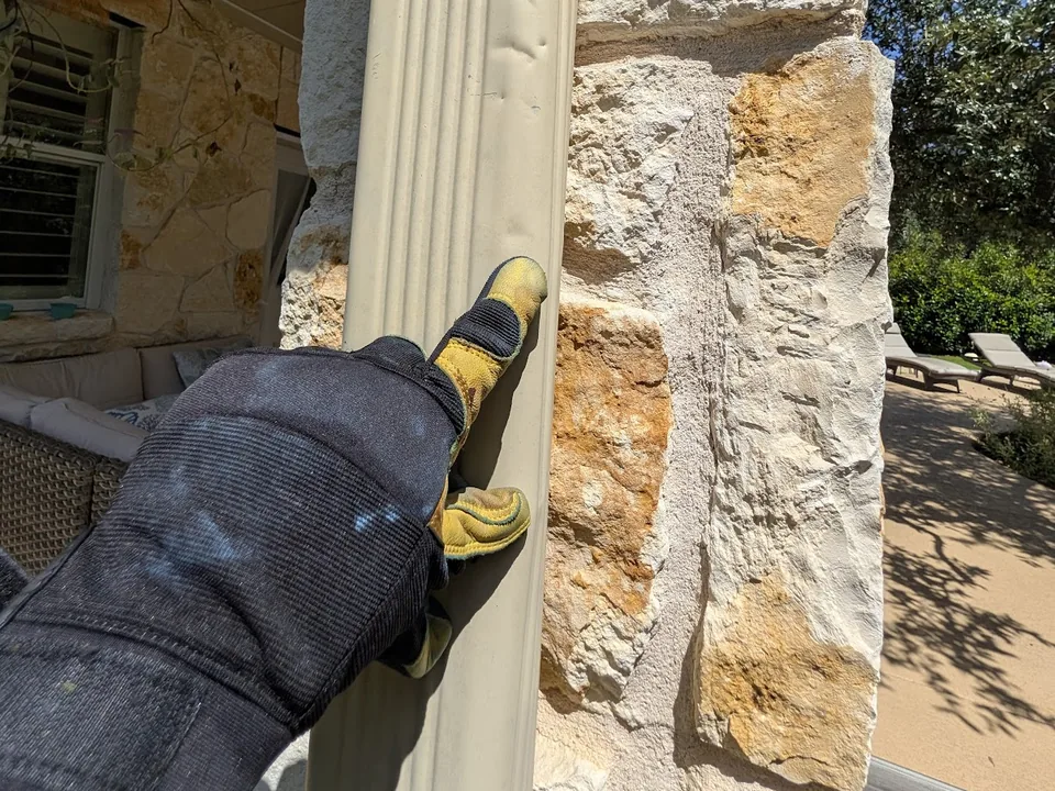 Gloved hand pointing at a dent in a tan downspout on a stone column