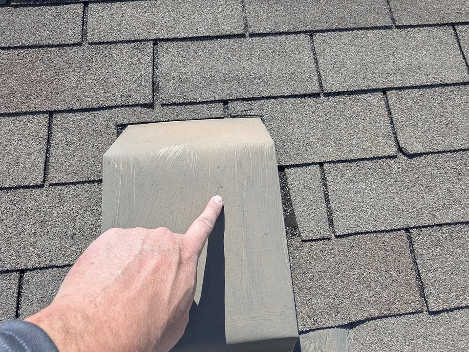 Hand pointing at a painted slant-back roof vent on a shingle roof